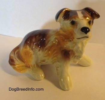 The front right side of a white with brown porcelain Scotch Collie dog figurine in a sitting position. The figurine has black circles for eyes and it has a line for a mouth.
