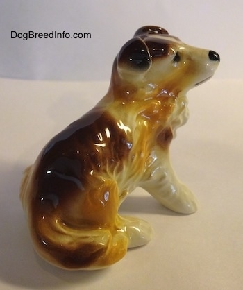 The back right side of a white with brown porcelain figurine of a Scotch Collie dog sitting. The figurine has small flopped over ears.