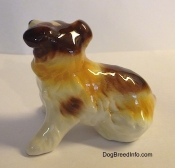 The left side of a white with brown porcelain figurine of a Scotch Collie sitting. The figurine is glossy.