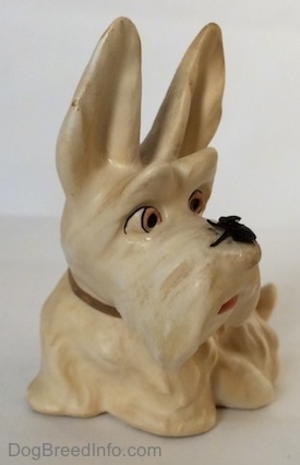 A white and cream figurine of a Scottish Terrier sitting. The figurine has a fly on its muzzle and its mouth painted open.