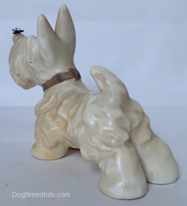 The back left side of a white and cream Scottish Terrier figurine with a fly on its nose. The figurine has fine hair details along its body.