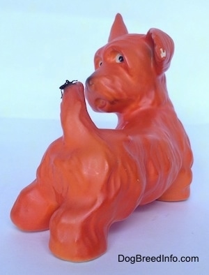 The back right side of a figurine of an orange with black Scottish Terrier. The figurine has white circles with black circles inside of it for eyes.