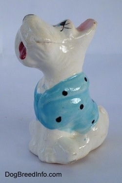 The right side of a white ceramic figurine of a Scottish Terrier sitting and it has a blue with black shirt on. The figurines legs are attached.