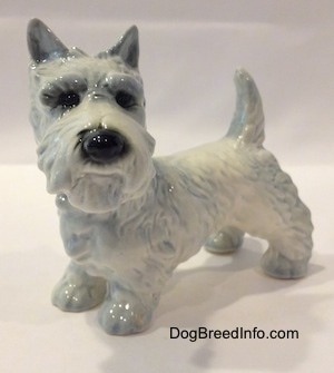 The front left side of a white with blue highlights Scottish Terrier figurine. The figurine has its ears in the air.