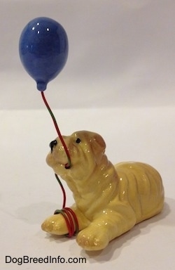 The front left side of a Shar-Pei figurine in a lying position with a blue balloon in its mouth.