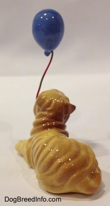 The back left side of a Shar-Pei figurine in a lying position. The figurine has a blue balloon in its mouth. The ears of the figurine are hard to differentiate from its head.