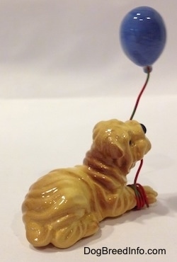 The back right side of a figurine of a brown and tan Shar-Pei in a lying posiditon with a blue balloon string in tis mouth. The figurine has a tail that is hard to see.