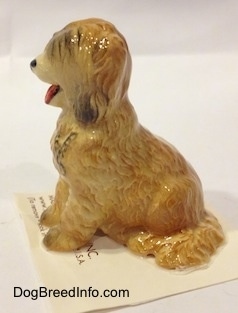 The left side of a figurine of a tan with brown Sheepdog that is in a sitting pose. The figurine has a black chain painted onto its chest,