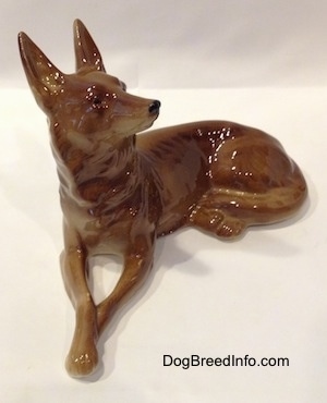 A brown with white Shepherd figurine in a lying pose. The figurine has tiny black circles for eyes.