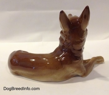 The right side of a figurine of a brown with white Shepherd lying. The figurine has alert ears.