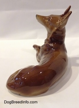 The back right side of a brown with white Shepherd lying figurine. The figurines tail is laying along its left side.