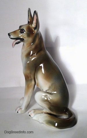 The left side of a figurine of a brown and white with black sitting German Shepherd. The figurine has a long tail that is layed to the left side of the figurine.