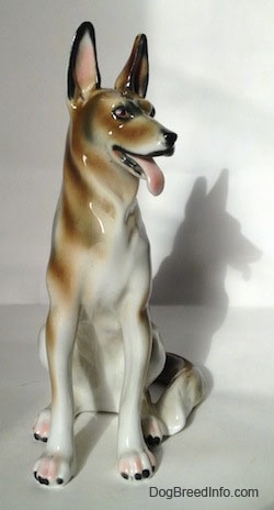 A white with brown and black figurine of a German Shepherd sitting. The figurine has black tipped nails.