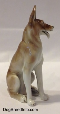 The front right side of a white and brown figurine of a German Shepherd sitting. The figurine has long white legs with black tipped nails.