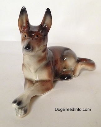 The front left side of a brown and white with black figurine of a German Shepherd lying figurine. The figurine has tiny black circles for eyes.