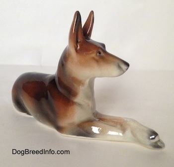 The front right side of a figurine of a brown and white with black German Shepherd lying.