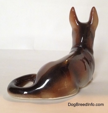 The back right side of a brown and white with black figurine of a lying German Shepherd. The tail of the figurine is long and wrapped around to the left side.