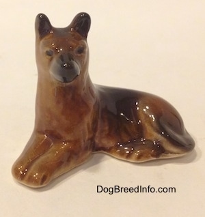 The front left side of a brown with black Shepherd figurine in a lying pose. The figurine has small black circles for eyes.