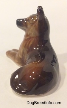 Tha back of a figurine of a brown with black Shepherd in a lying pose. The figurines tail is indistinguishable from its back side.