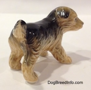 The back right side of a figurine of a black with tan German Shepherd puppy standing. The figurines ears are black tipped and flopped over.
