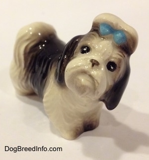 The front left side of a white with black figurine of a Shih Tzu with a blue bow in its hair. The figurine has black circles with white circles inside of it for eyes.
