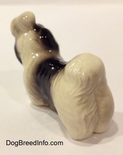 The back left side of a figurine of a white with black Shih Tzu with a blue bow in its hair. The figurine has a lump of hair on its head.