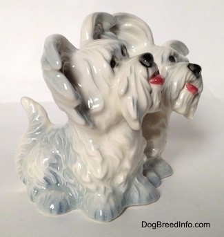 The front right side of a Siamese twin silver platinum figurine of a sitting Skye Terrier. The figurines have black circles for eyes.