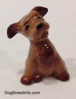 A brown Terrier puppy figurine in a sitting pose. The figurines ears are lifted up and its head is tilted to the left.