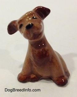 The front left side of a figurine of a brown Terrier puppy in a sitting pose. The figurine has black circles for eyes and a nose.