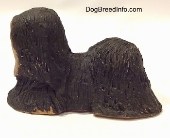 The left side of a figurine of a black with tan Tibetan Terrier standing. The figurine has short legs.