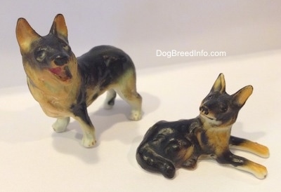 The front left side of a standing porcelain figurine of a black and tan German Shepherd standing with its mouth open figurine. Next to it is the right side of a laying porcelain of a black and tan German Shepherd figurine.
