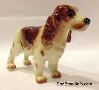 The front right side of a figurine of a red and white standing Welsh Springer Spaniel. The figurine has a black nose.