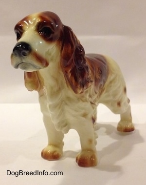 The front left side of an orange and white figurine of a standing Welsh Springer Spaniel. The figurine has hair details on its chest.