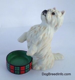 The front left side of a West Highland White Terrier figurine in a begging pose next to an empty dish. The figurine has short standing ears.
