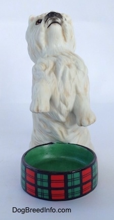 A figurine of a West Highland White Terrier in a begging pose with an empty dish in front of it. The figurine has short legs.