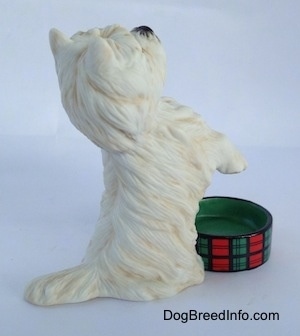 The right side of a West Highland White Terrier in a begging pose with an empty dish in front of it figurine. The figurine has a short tail.