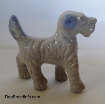 The right side of a figurine of a porcelain white with blue Wire Fox Terrier. The figurie has the word 'JAPAN' engraved on its side.