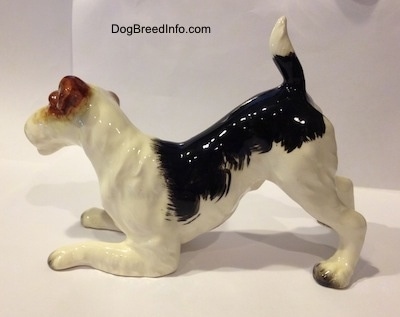 The left side of a porcelain figurine of a black and white with brown Wire Fox terrier in a play bow pose. The figurine has its tail in the air.