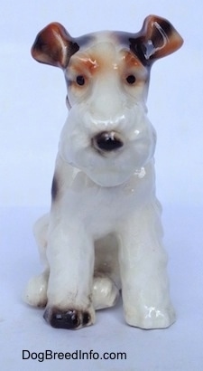 A white with black and brown figurine of a Wire Fox Terrier sitting. The figurine has medium length legs.