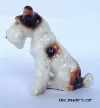 The left side of a white with black and brown figurine of a Wire Fox Terrier sitting figurine. The figurine has a black and brown spot aboce its leg.