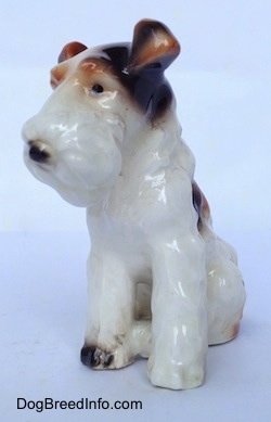 The front left side of a figurine of a white with black and brown sitting Wire Fox Terrier. The ears of the figurine are flopped over.