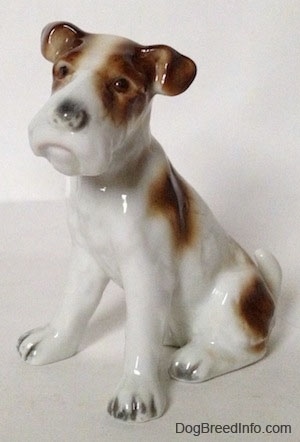 A white with brown figurine of a Wire Fox Terrier that is sitting. The figurine has detailed brown with black eyes.