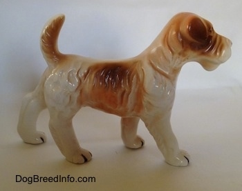 The right side of a white and tan figurine of a standing Wire Fox Terrier. The figurine has an arched up tan tail.