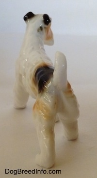 The back left side of a figurine of a bone china white with black and tan Wire Fox Terrier. The figurines tail is arched in the air.