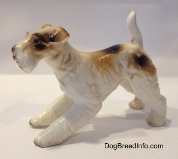 The left side of a ceramic white with tan and black figurine of a Wire Fox Terrier. The figurine has fine hair details along its body.