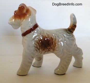 The left side of a porcelain white with tan standing Wire Fox Terrier figurine. The figurine has fine hair details along its body and a big brown spot on its side.