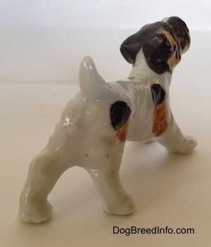 The back right side of a bone china white with black and brown Wire Fox Terrier figurine in a play bow pose. The figurine has a short tail that is arched in the air.