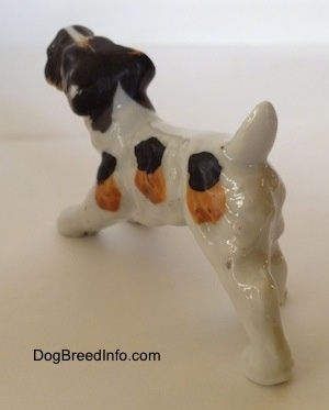 The back left side of a figurine of a bone china white with black and brown Wire Fox Terrier in a play bow pose. The figurine is glossy.
