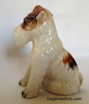 The left side of a figurine of a ceramic white with black and brown Wire Fox Terrier in a sitting pose. The figurine has brown flopped over ears.