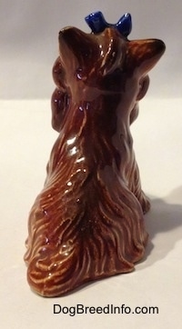 The back of a figurine of a brown with tan sitting Yorkshire Terrier with a bluw bow on its head. the ears of the figurine are in the air.
